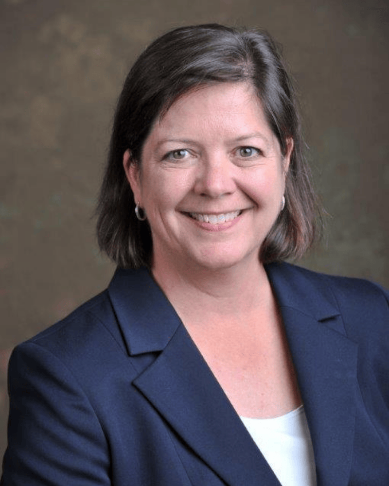 Professional headshot of a woman with short brown hair wearing a navy blazer and white top, smiling against a neutral studio background.